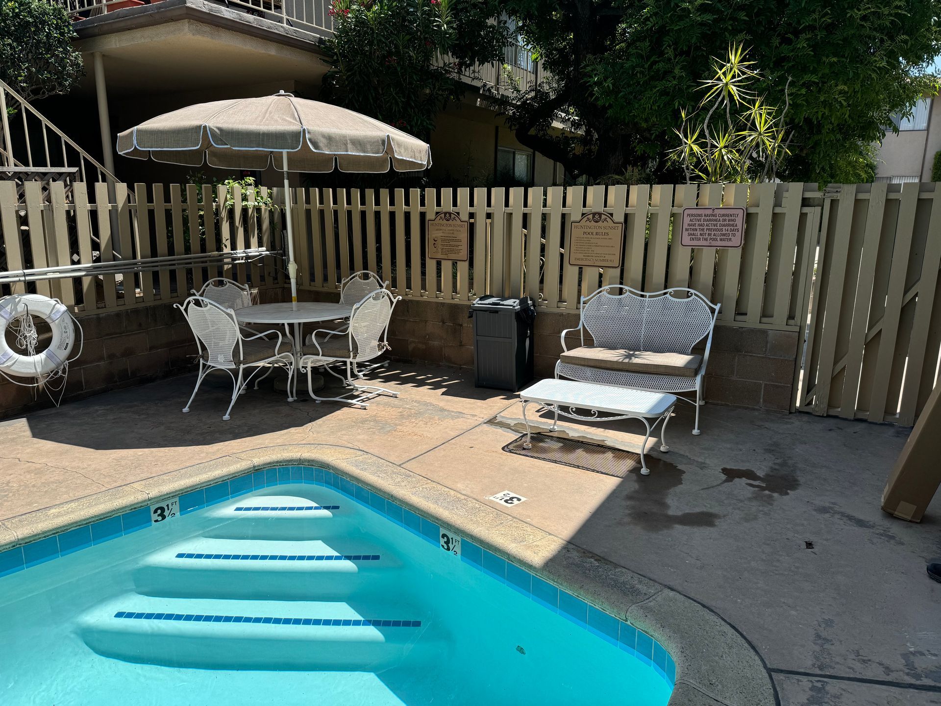 A poolside seating area with a table, umbrella, and bench on a concrete patio next to swimming pool steps.