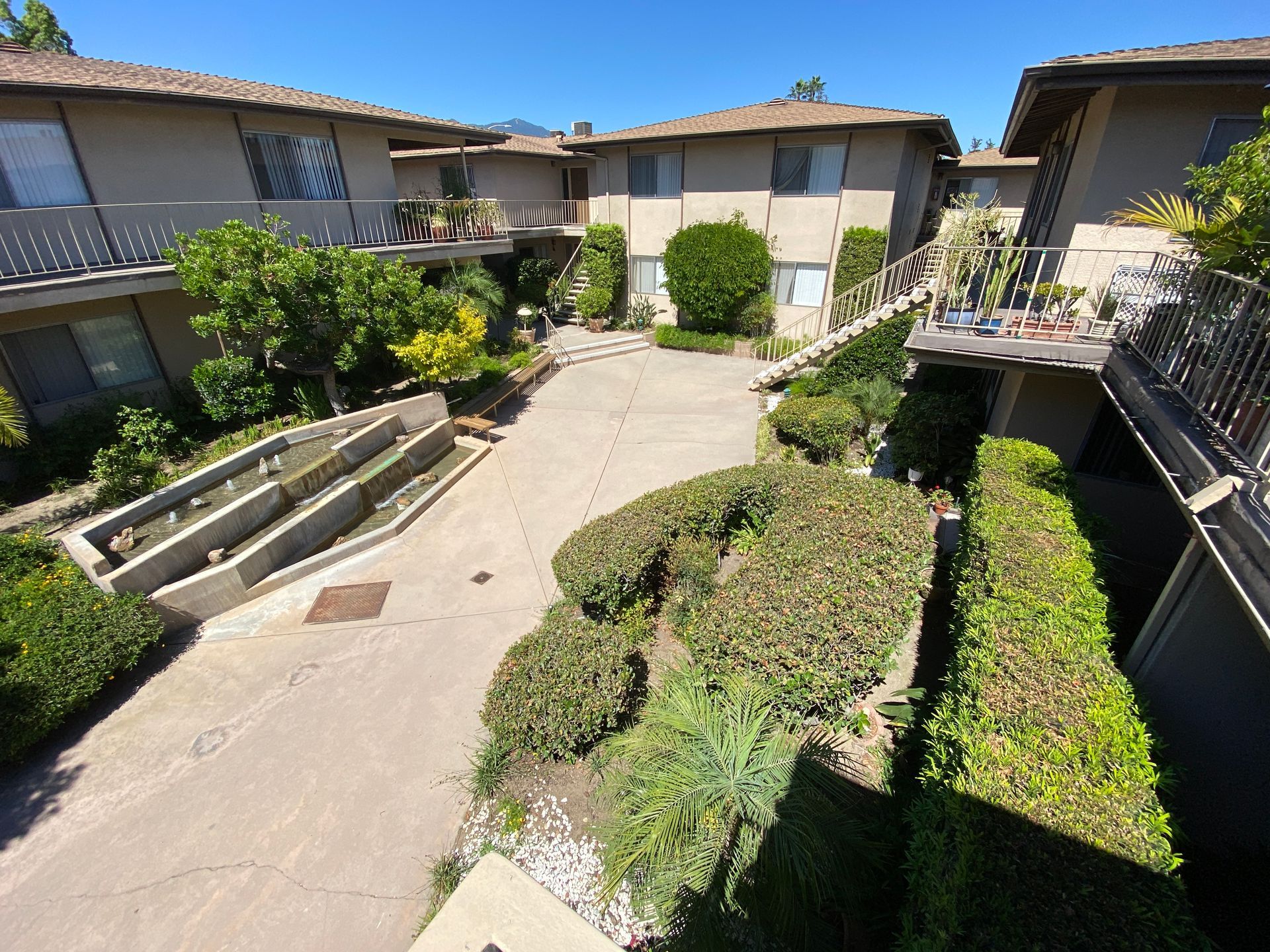 A sunny courtyard at a two-story apartment complex featuring concrete pathways, manicured shrubs, and tiered planters.