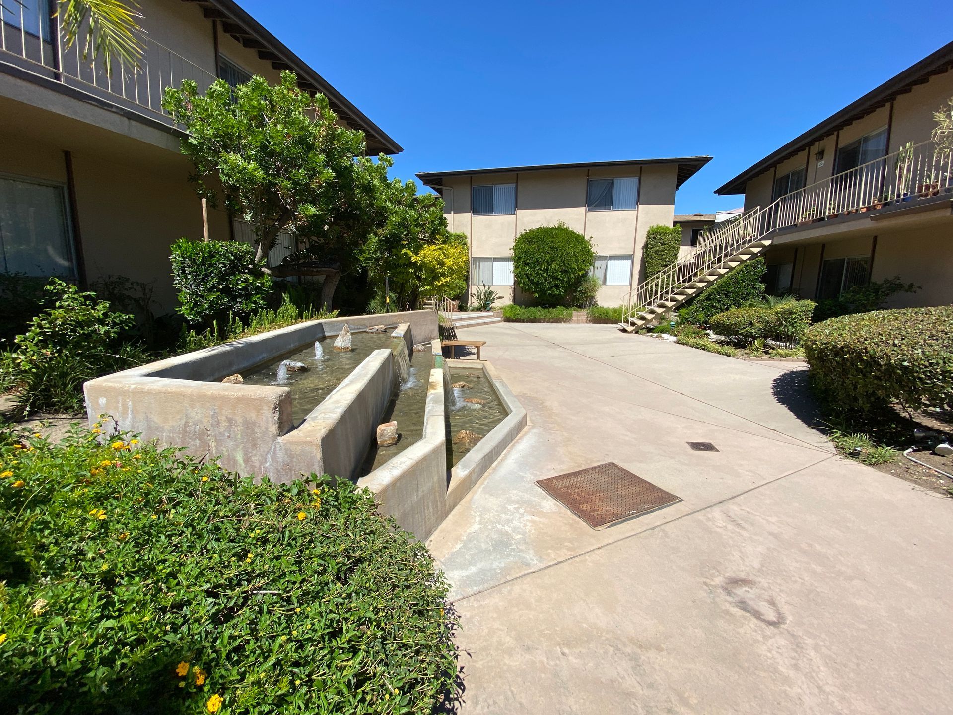 A courtyard featuring a tiered concrete fountain, lush greenery, and apartment buildings under a clear blue sky.