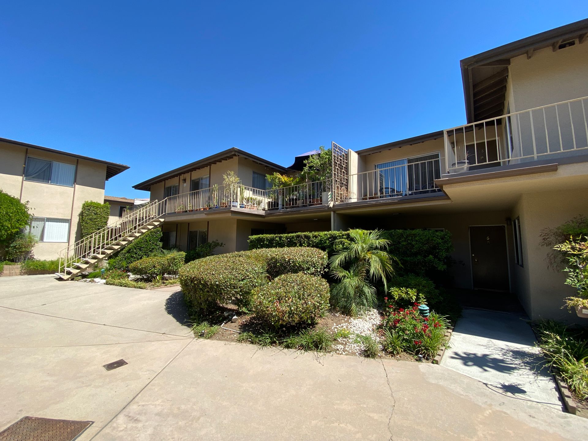 A tan, two-story apartment complex with wooden balconies and stairs, surrounded by shrubs and trees under a clear sky.