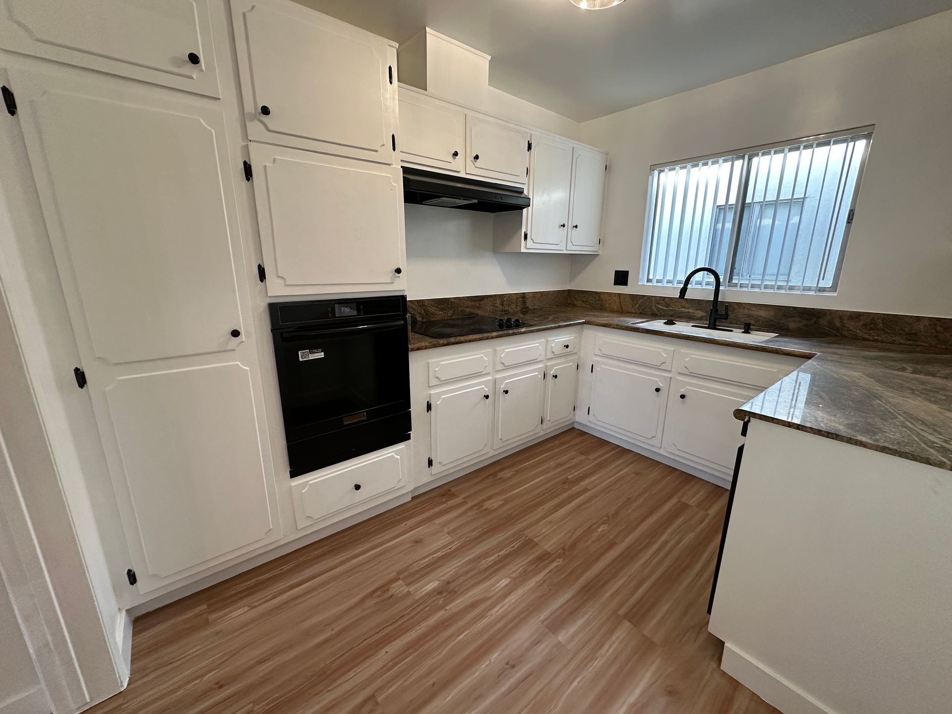 A bright kitchen with white cabinets, dark countertops, a black wall oven, a sink by a window, and wood-look flooring.