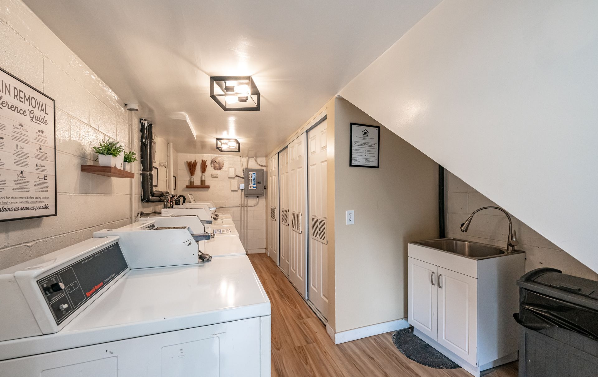 A laundry room with white appliances, wooden flooring, recessed lighting, and a utility sink under a stairwell.