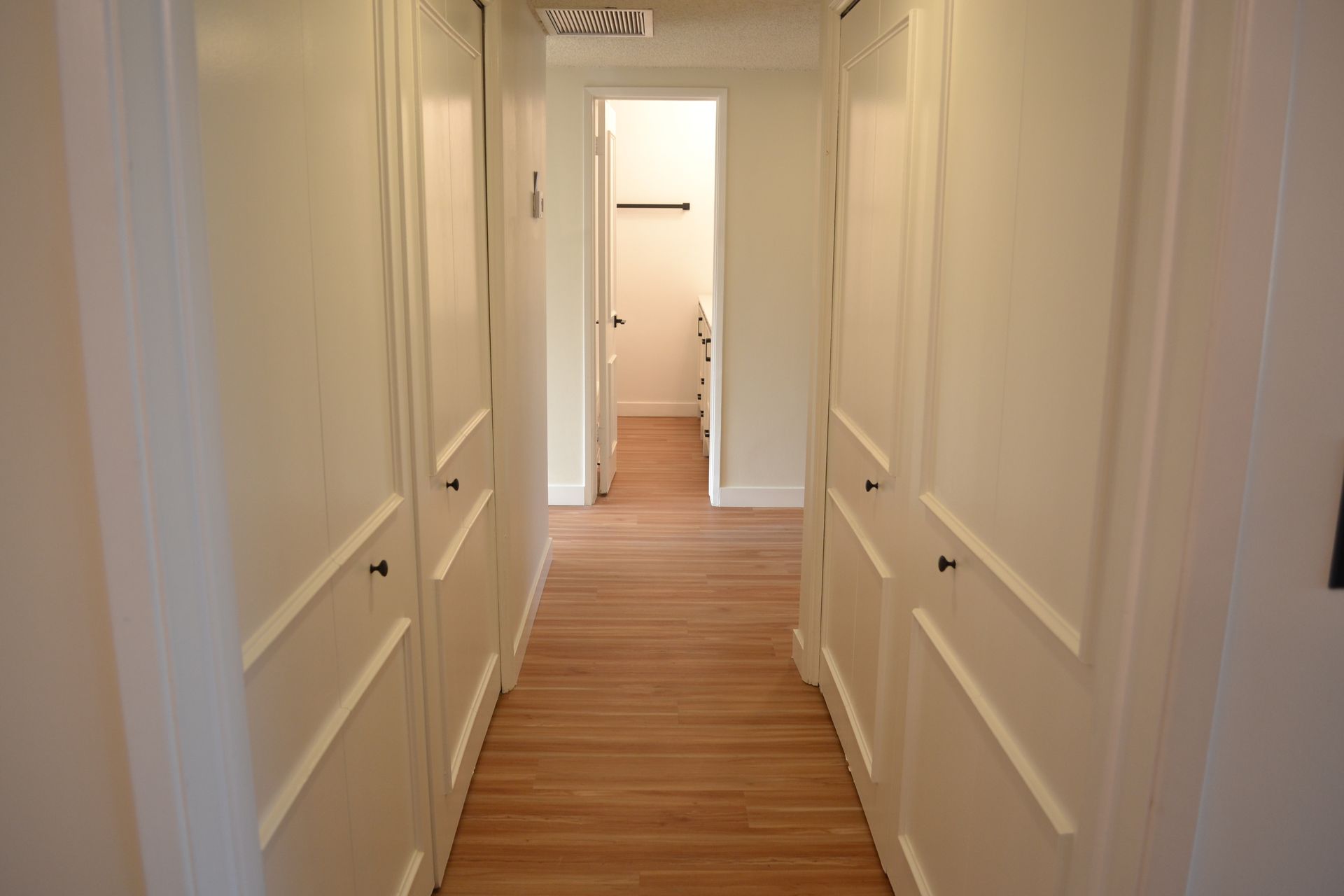 A brightly lit hallway with white walls and wood-look flooring, lined with white paneled closet doors on both sides.