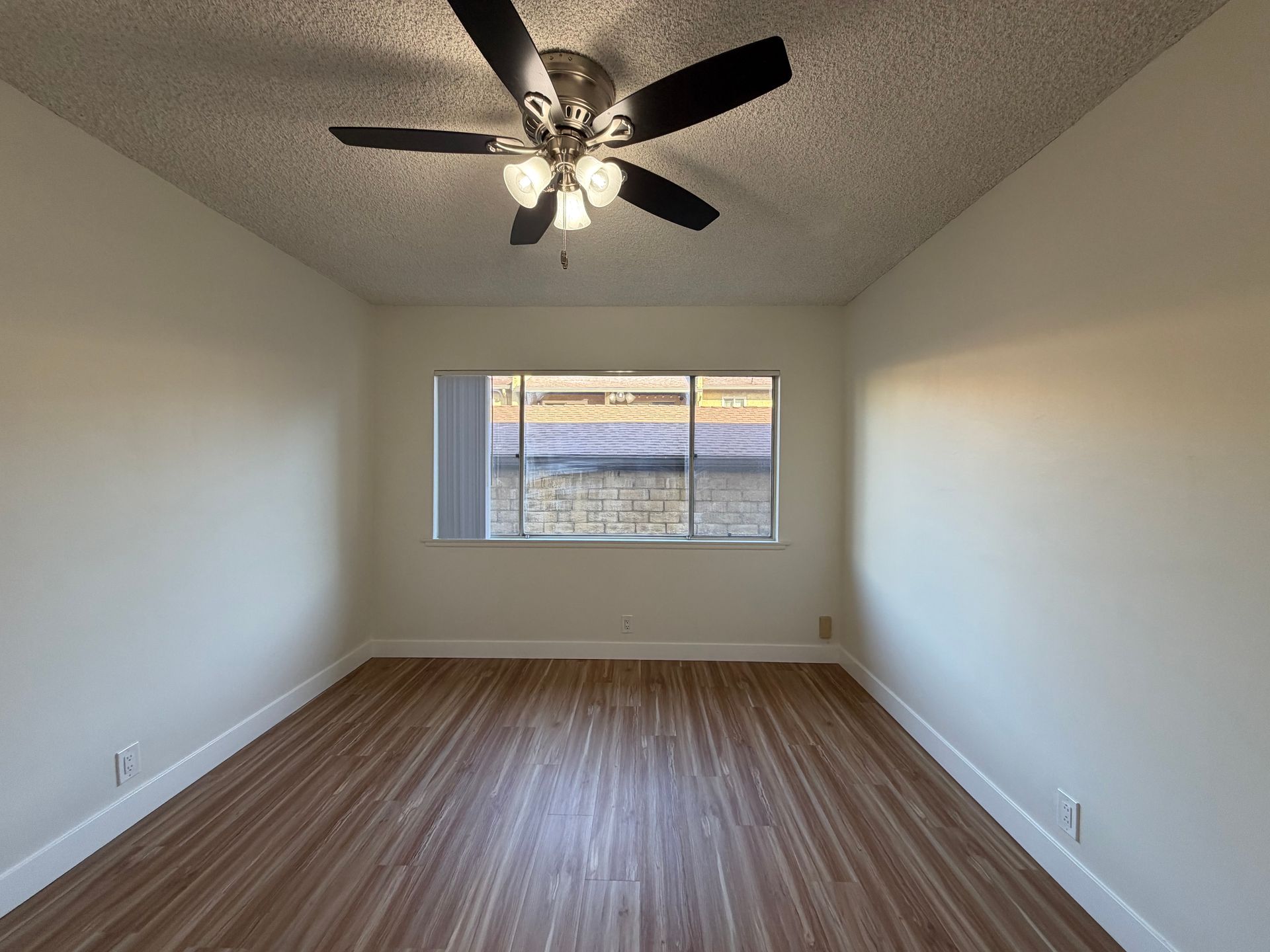 Empty room with off-white walls, wood-look flooring, a ceiling fan, and a window with a view of a street.