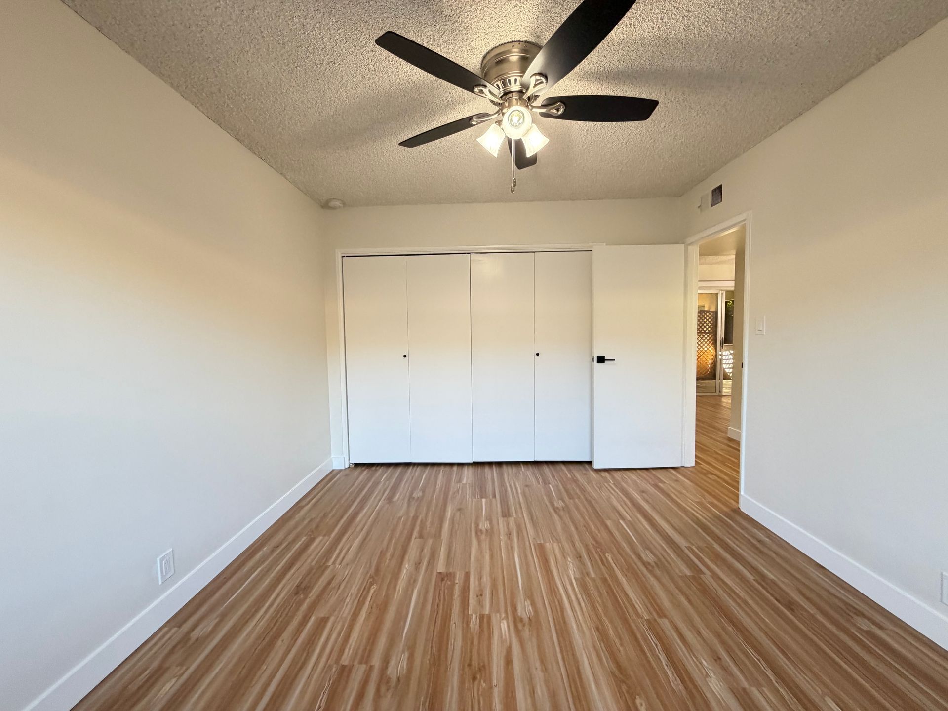 An empty bedroom with light-colored walls, wood-look flooring, a ceiling fan, and a wall of white closet doors.