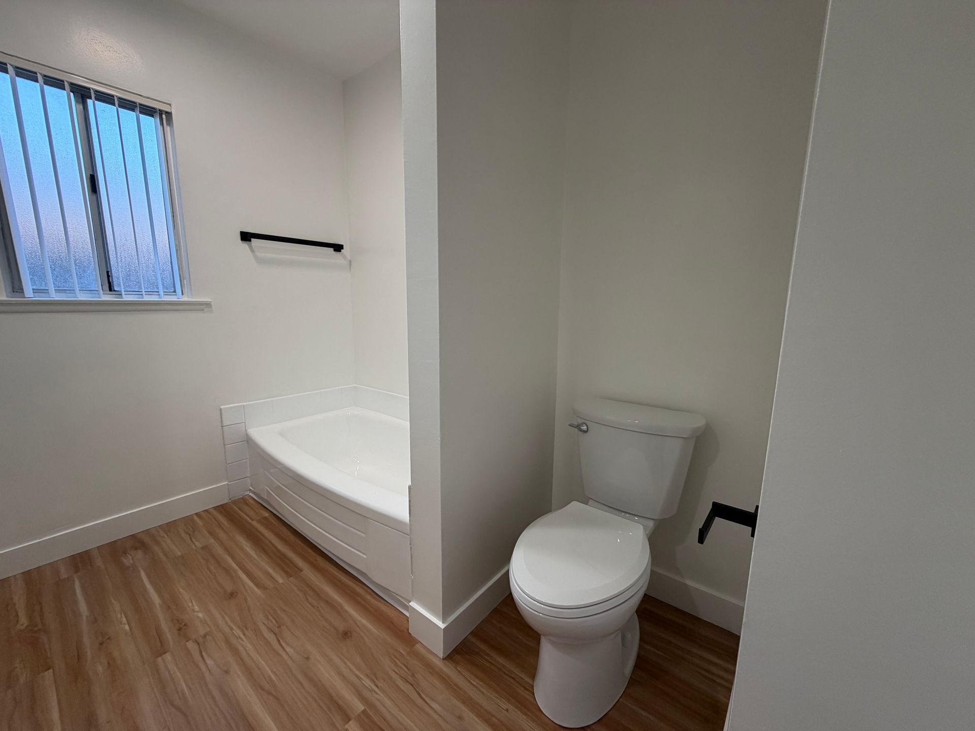 A bathroom featuring a white toilet, a built-in bathtub against a white wall, and light wood-tone flooring.