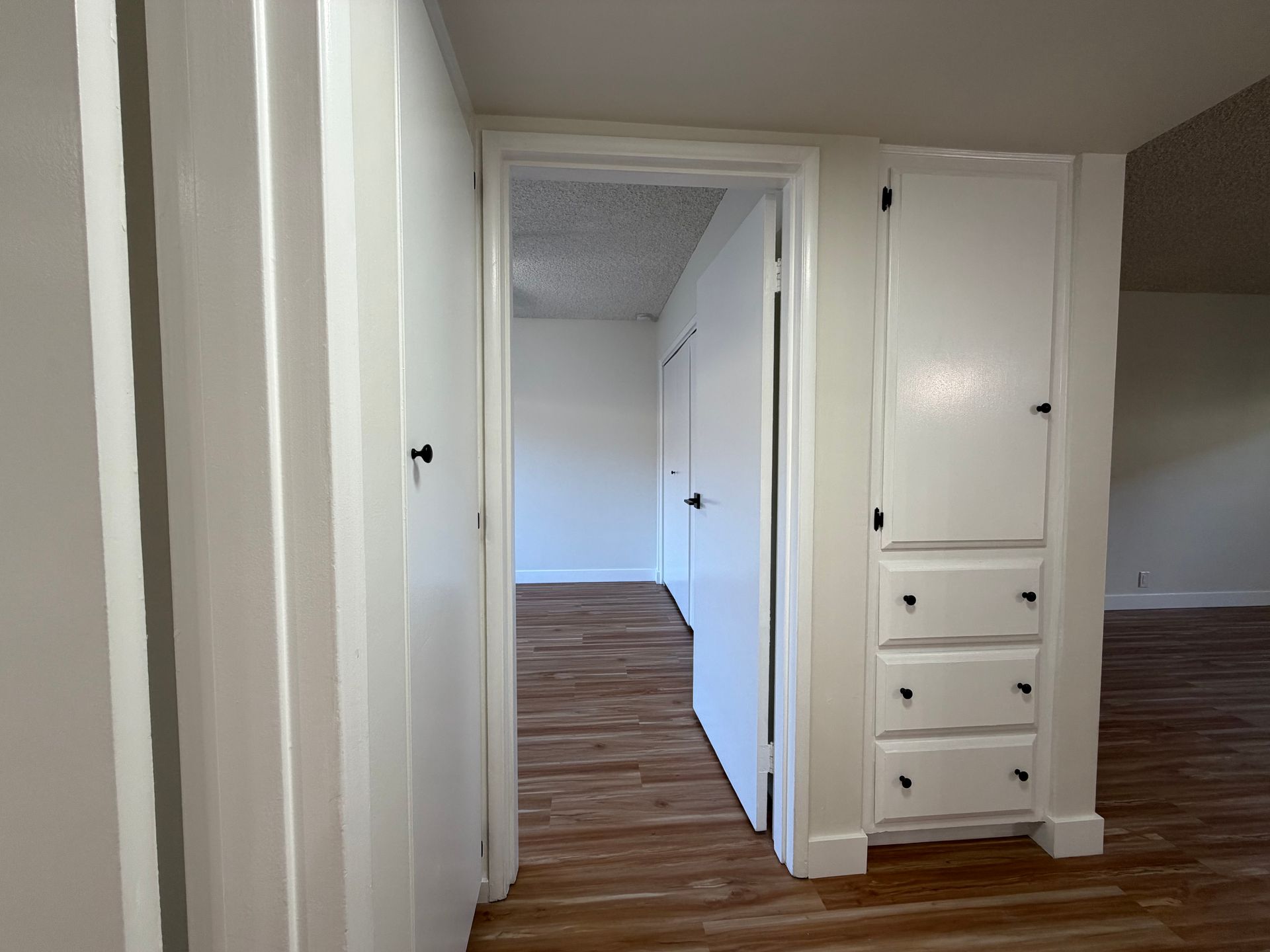 A hallway with wood-look flooring leads to an open room, featuring a white built-in cabinet with drawers on the right.