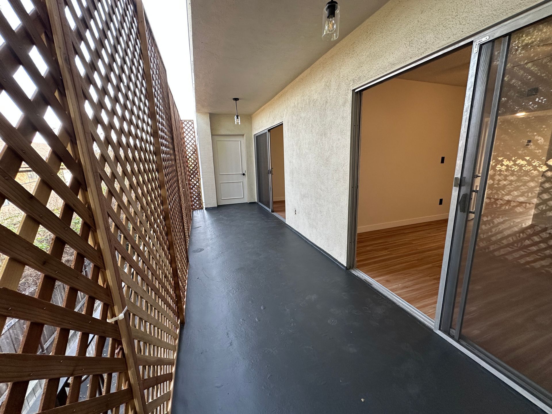 A balcony with dark flooring, a wooden lattice screen on the left, and a white sliding glass door leading to a room.