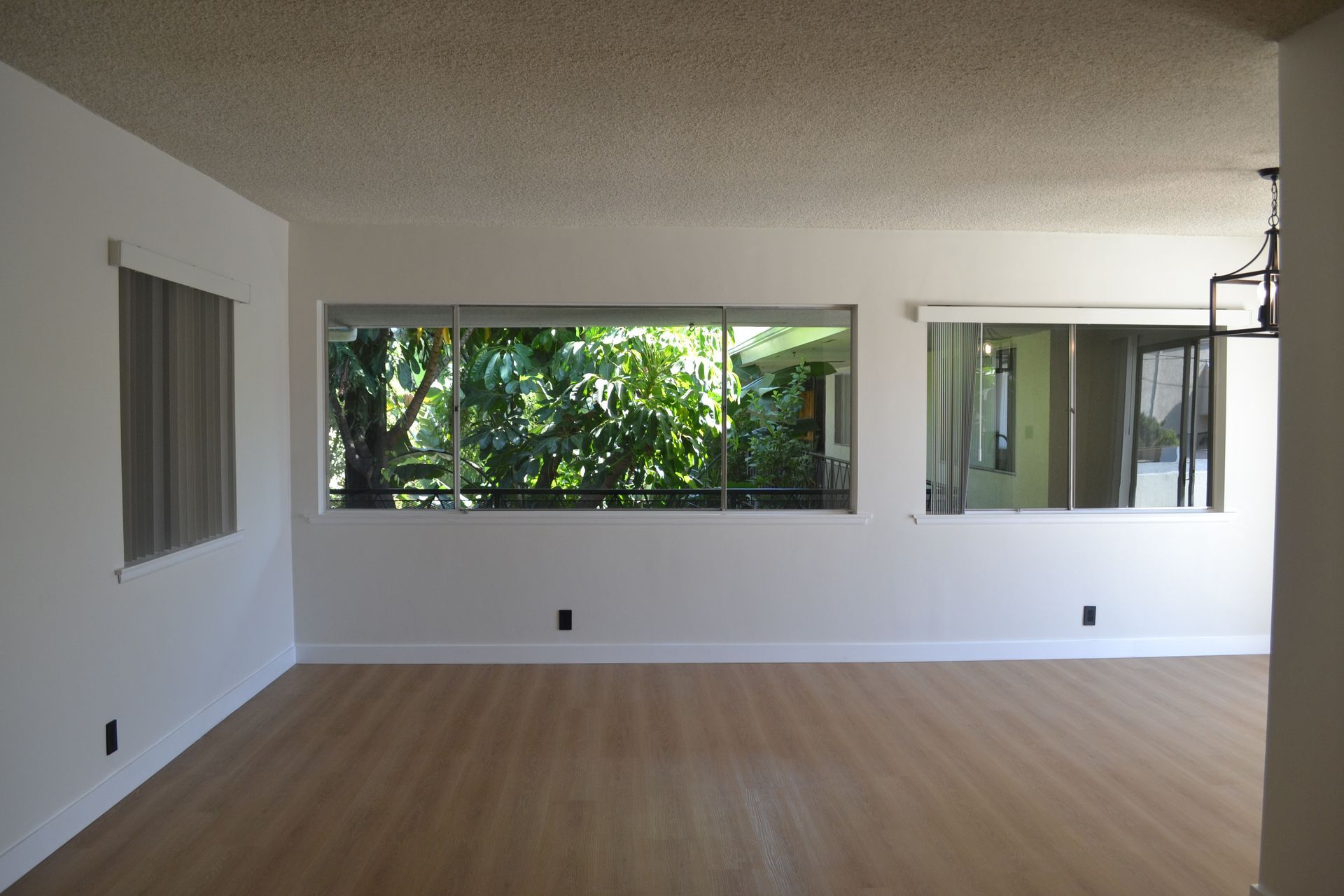 A bright, empty living room with wood-look flooring, white walls, a textured ceiling, and large windows with gray blinds.