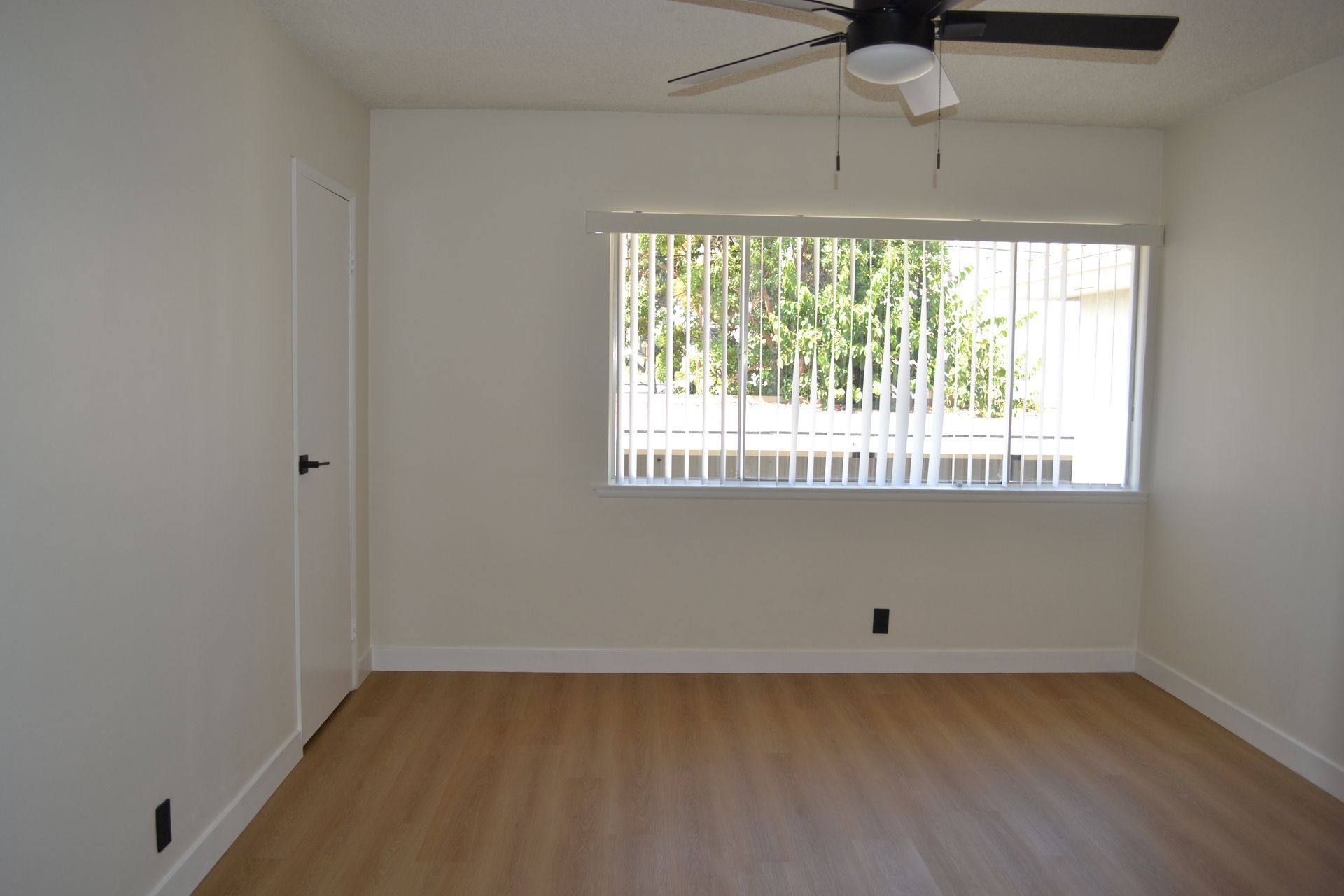 An empty room with light wood-look flooring, white walls, a closed white door, and a large window with vertical blinds.
