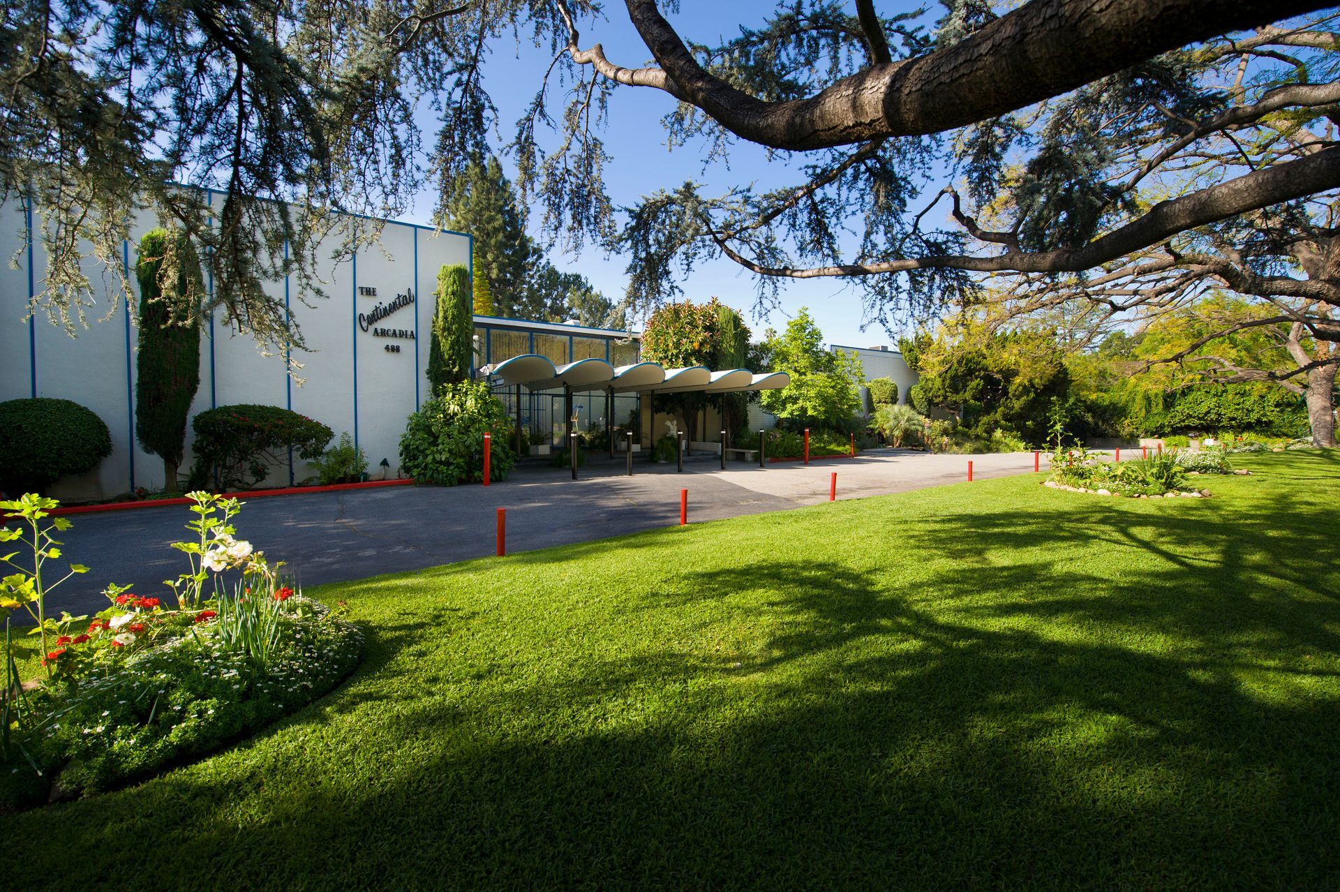 A modern building with light walls and a dark roof overhang, surrounded by manicured lawns, trees, and flower beds.