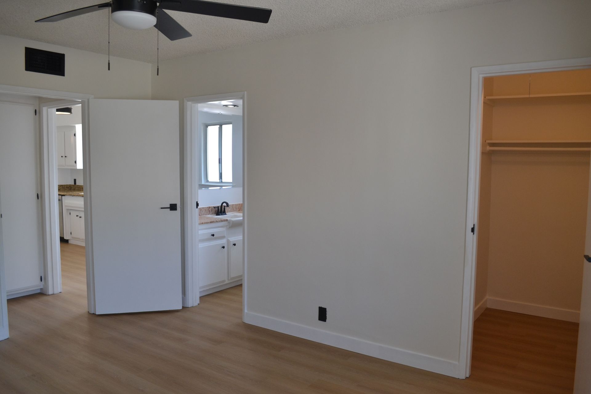 A view of a bright room with light wood floors, a ceiling fan, a walk-in closet, and open doorways to adjacent rooms.