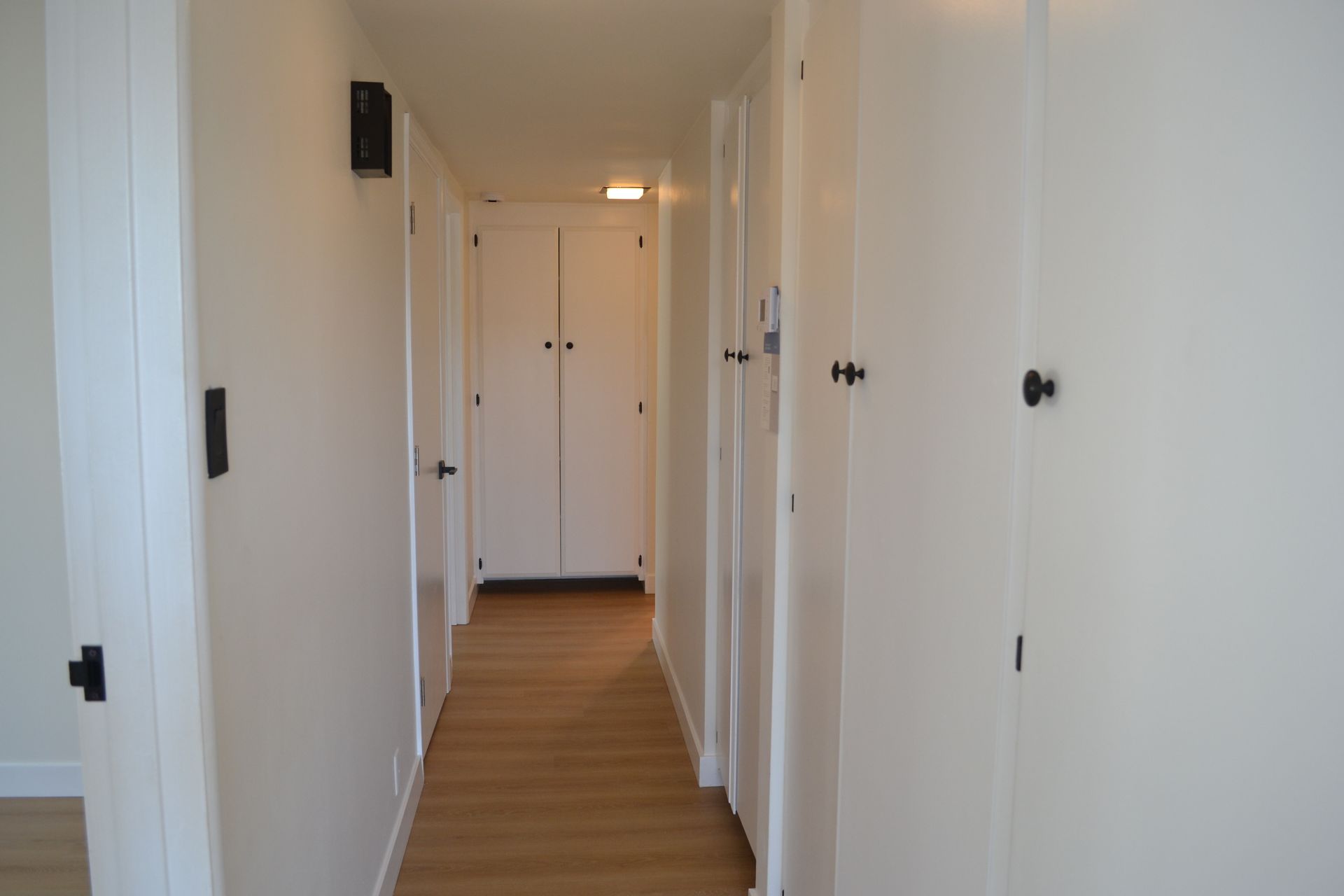 A white hallway with light wood flooring, featuring several white cabinet doors and a single door at the far end.