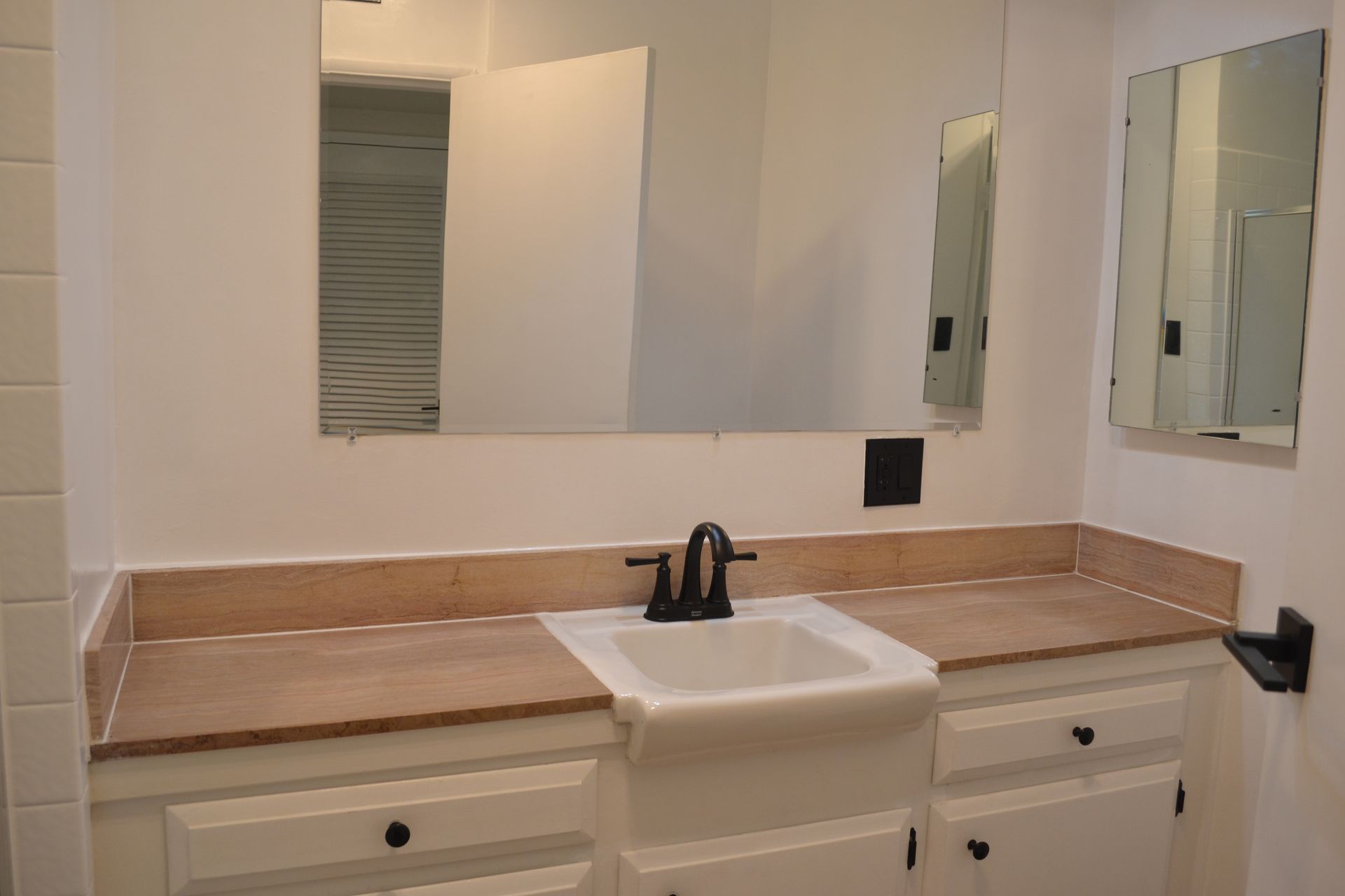 A bathroom vanity with a white farmhouse sink, tan countertop, white cabinets, and mirrors on a plain white wall.
