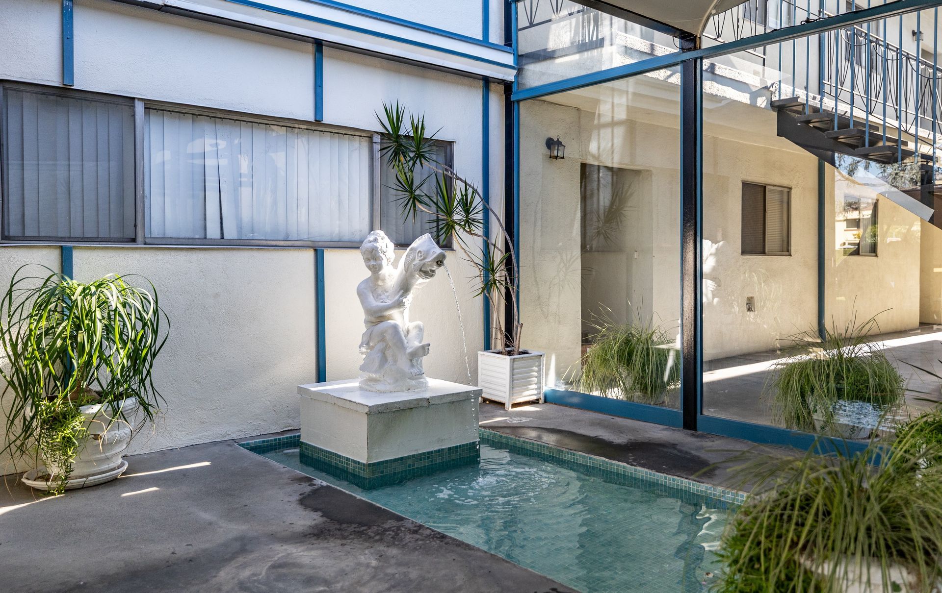 A white statue of a person sits on a pedestal in a small fountain, surrounded by potted plants in a paved courtyard.