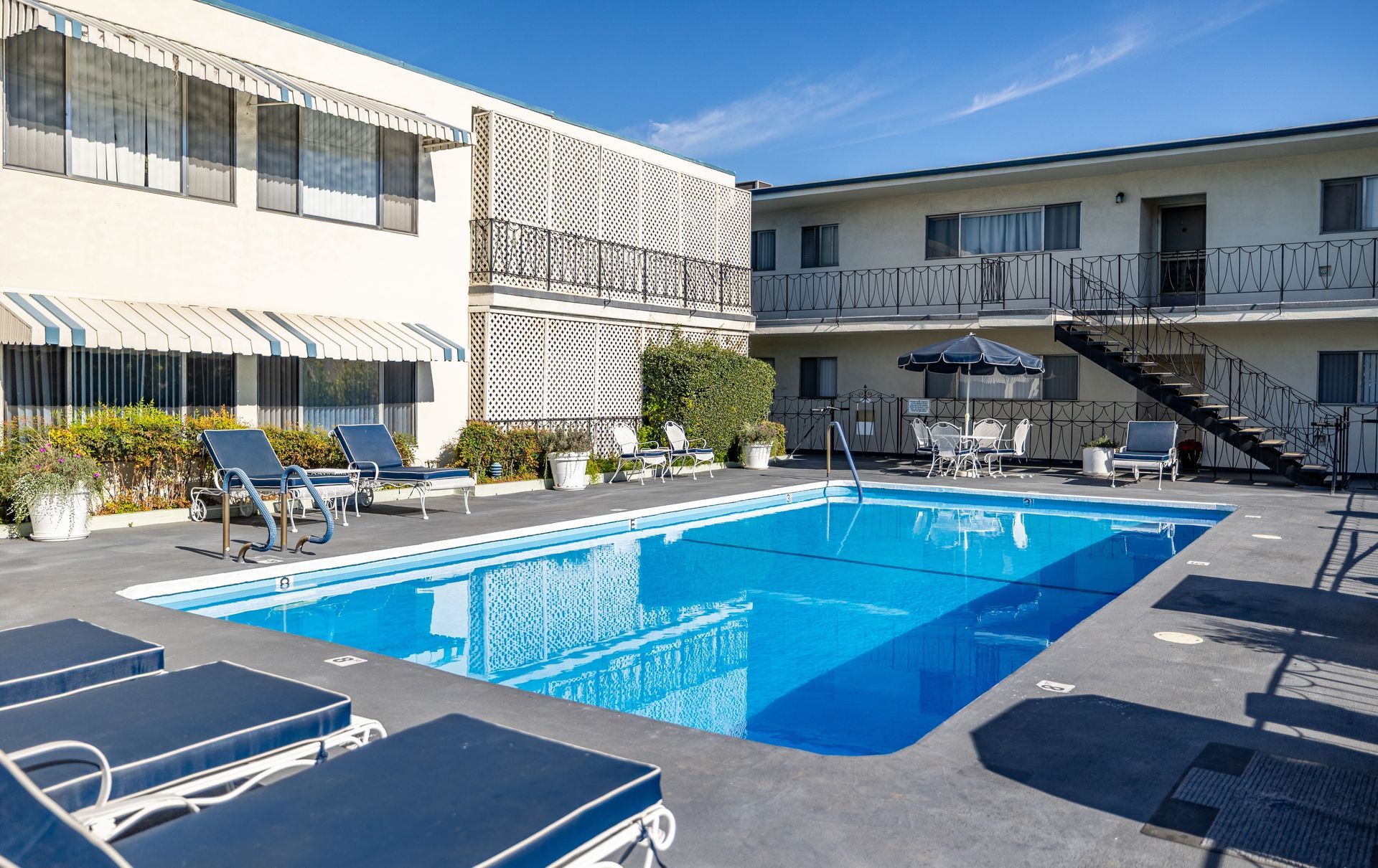 A swimming pool at a two-story apartment complex with blue lounge chairs on the concrete deck under a clear blue sky.