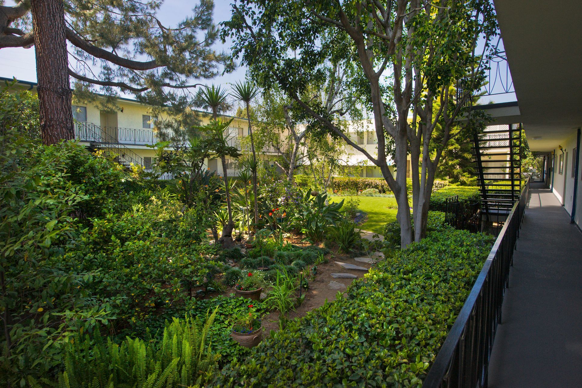 A multi-story apartment complex courtyard featuring lush green foliage, trees, and a walkway with a black metal railing.