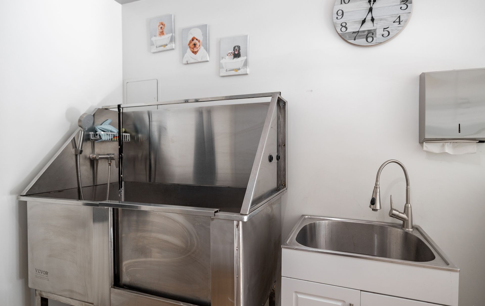 A stainless steel dog grooming tub and a matching basin sink in a clean, white room with wall art and a large clock.