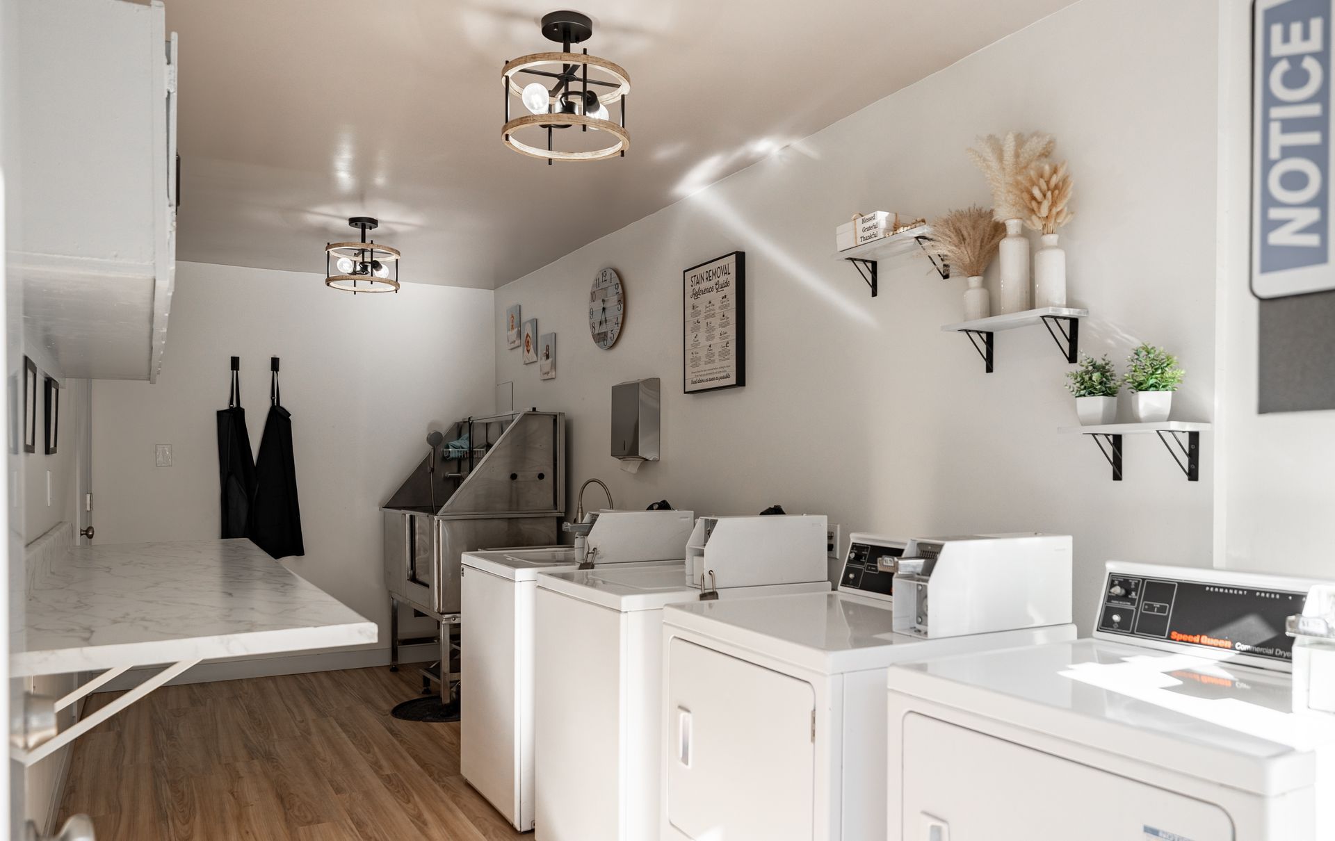 A laundry room featuring white washing machines, a stainless steel pet wash tub, wall-mounted shelves, and black lighting.