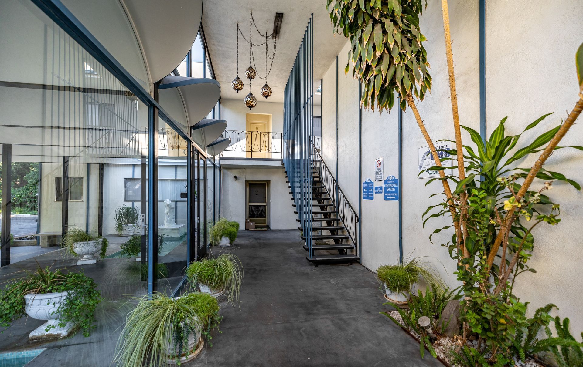 A brightly lit atrium with a central staircase, potted plants, hanging lights, and large windows looking outside.