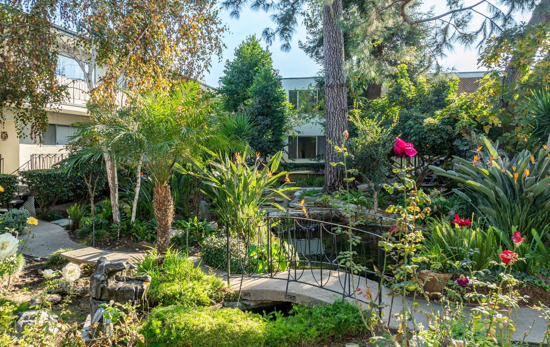 A sunny, lush garden courtyard featuring a small bridge, vibrant green foliage, and blooming red roses.