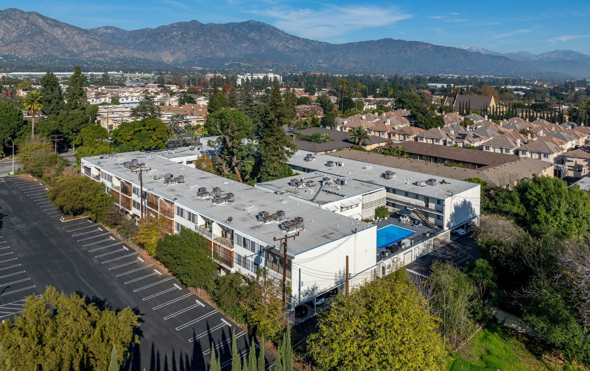 An aerial view of a multi-story apartment complex with a swimming pool, parking lot, and mountains in the distance.