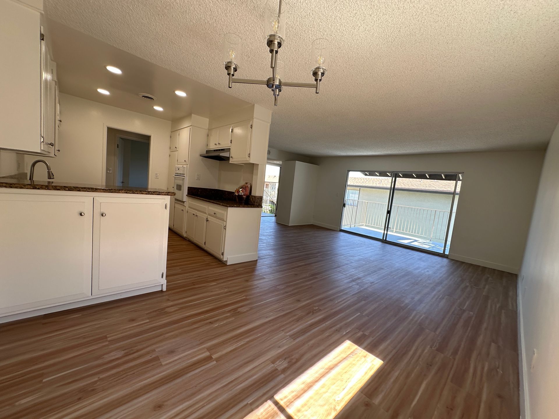 A bright, open-concept kitchen and living area featuring white cabinets, wood-look flooring, and a sliding glass door.