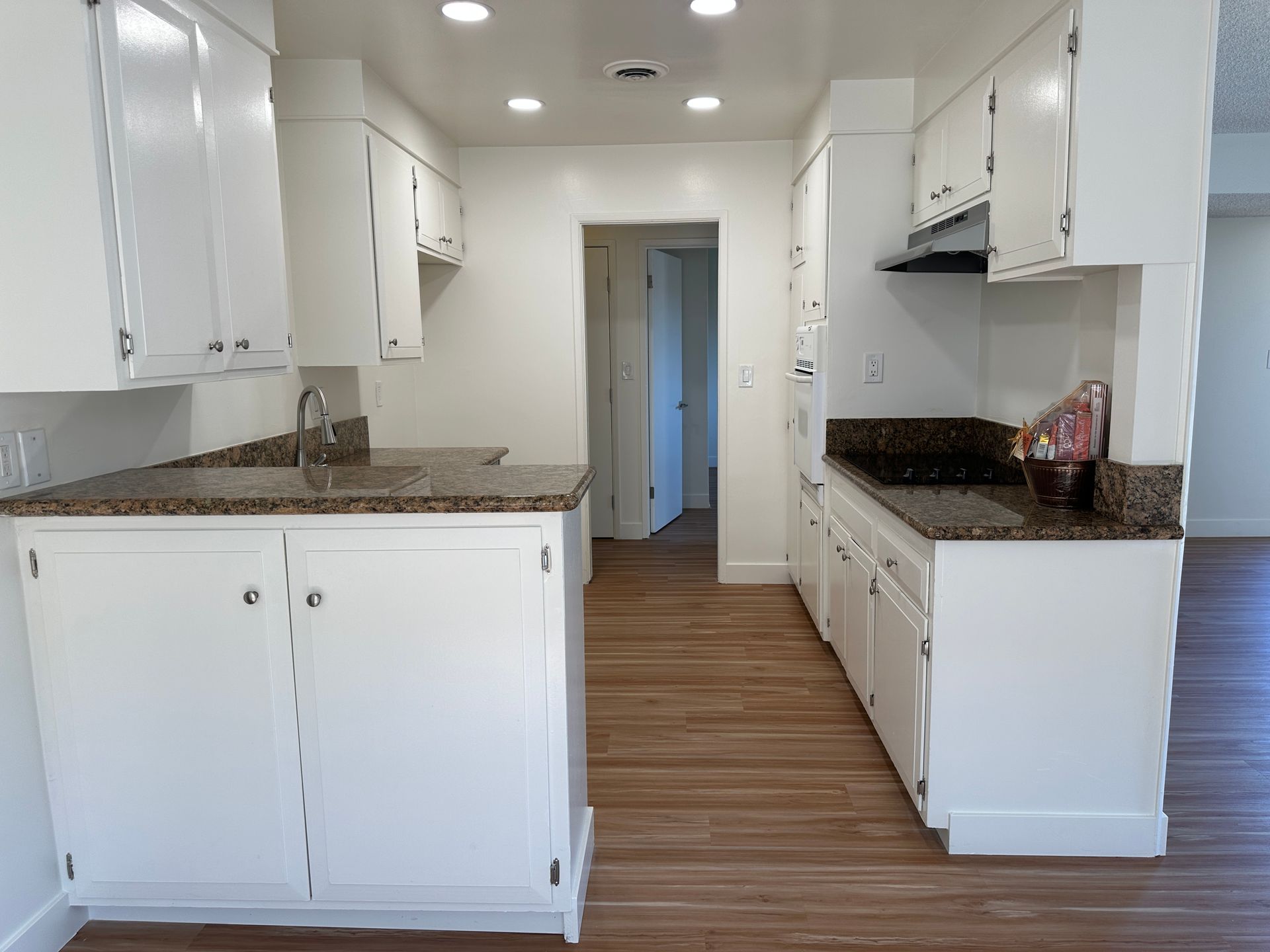 A kitchen with white cabinets, dark granite countertops, and light wood-tone flooring, viewed from an open doorway.