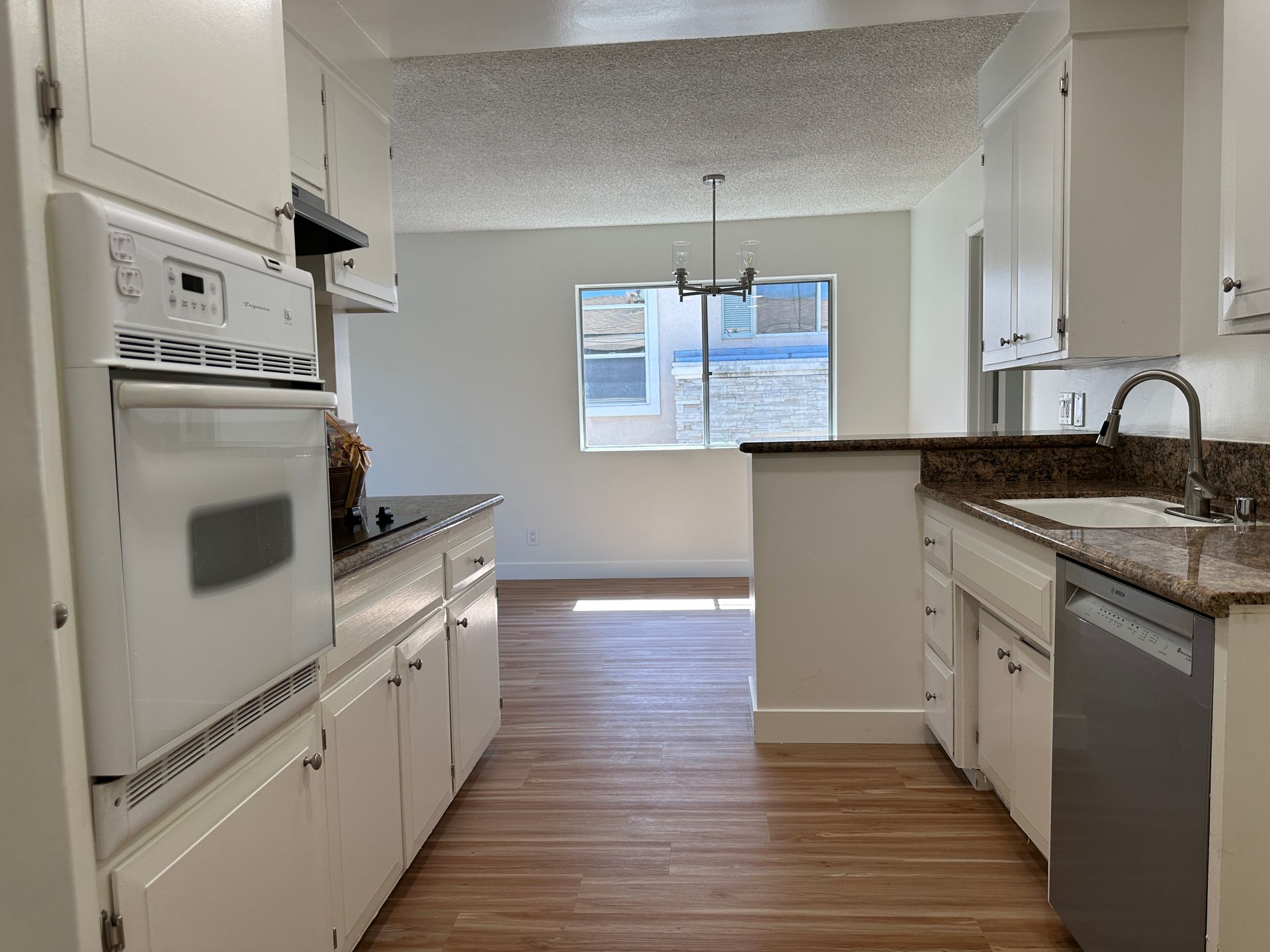 A galley-style kitchen with white cabinets, dark countertops, a wall oven, and a dishwasher, leading to a dining area.