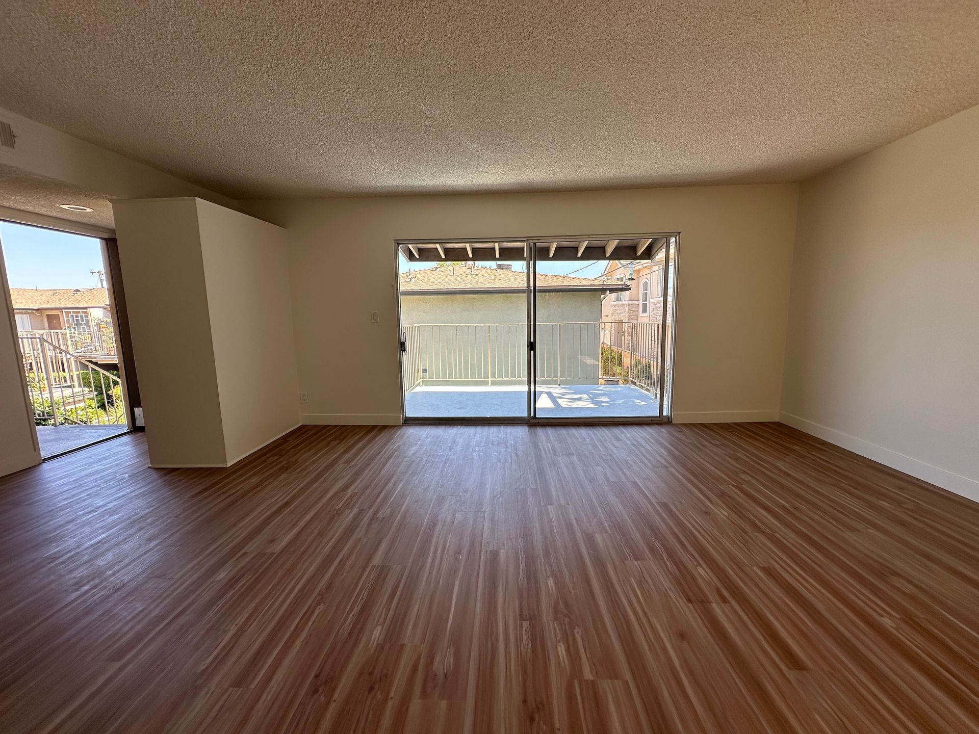 An empty room with light wood-look flooring, beige walls, a popcorn ceiling, and two sliding glass doors leading outside.