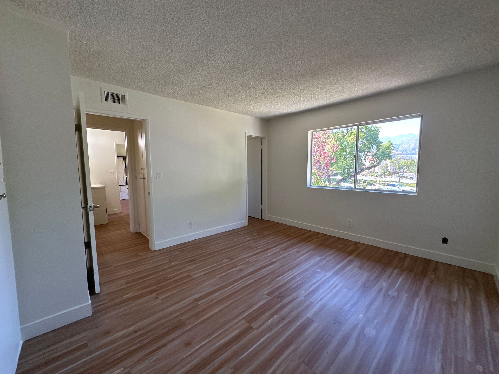 Empty room with light wood-style flooring, white walls, a textured ceiling, and a window looking out onto green trees.