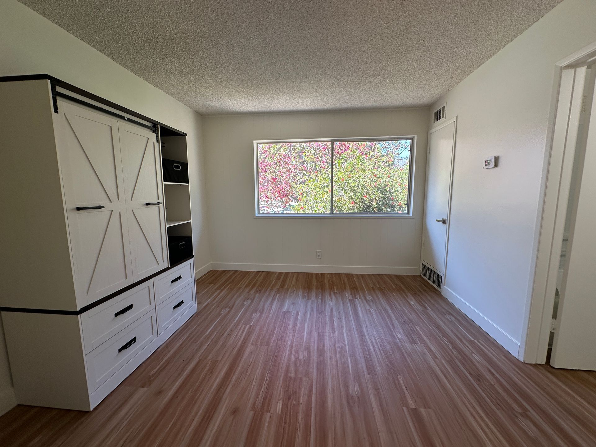 An empty bedroom with wood-look flooring, a large white cabinet with storage drawers, and a window viewing trees.