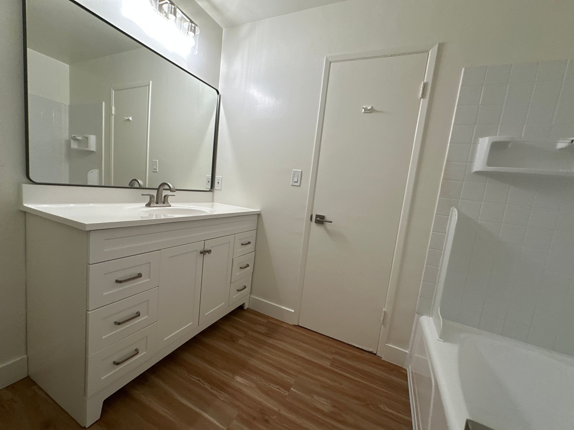 A modern bathroom with a white vanity, a large rectangular mirror, and a white door, featuring light wood-tone flooring.