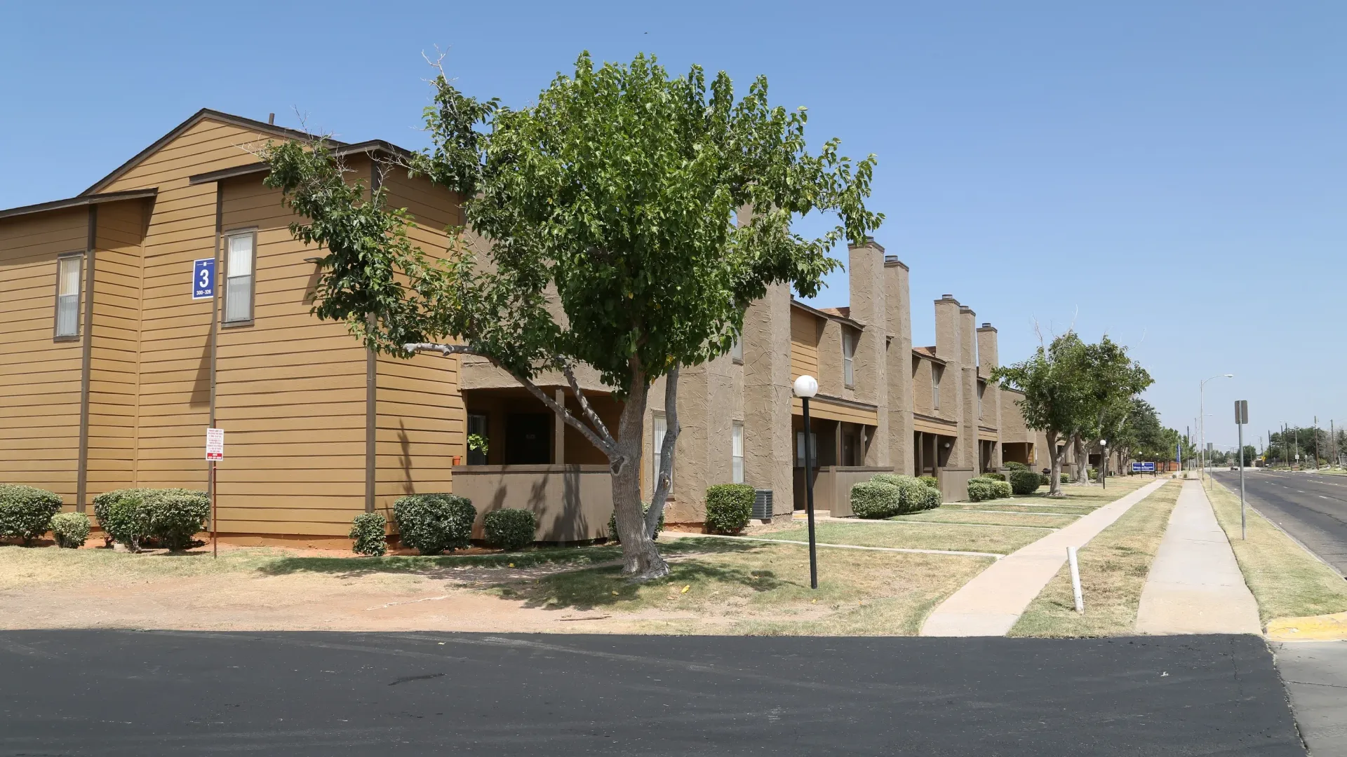 Apartment complex with brick exteriors and small trees lining the sidewalk next to a road under a blue sky.