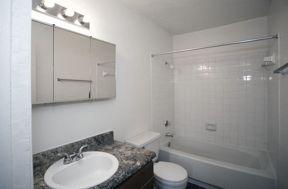 Apartment bathroom with a granite vanity, mirror cabinet, toilet, and white tiled tub/shower.