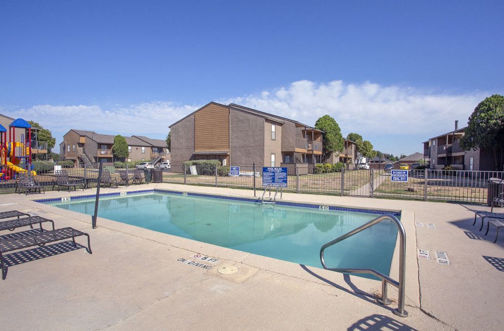 Pool and apartment buildings on a sunny day. Lounge chairs sit poolside. Playground in the background.