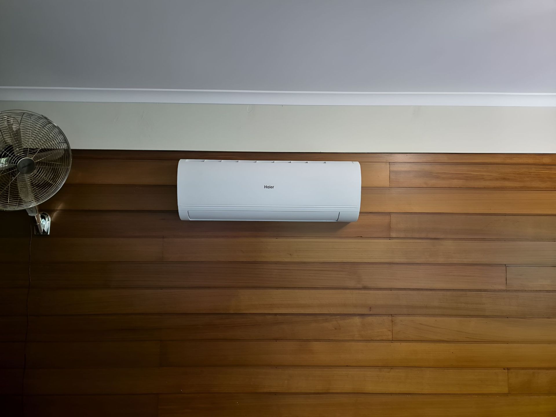 A White Air Conditioner is Mounted on a Wooden Wall Next to a Fan — Our Sparky In Urraween, QLD