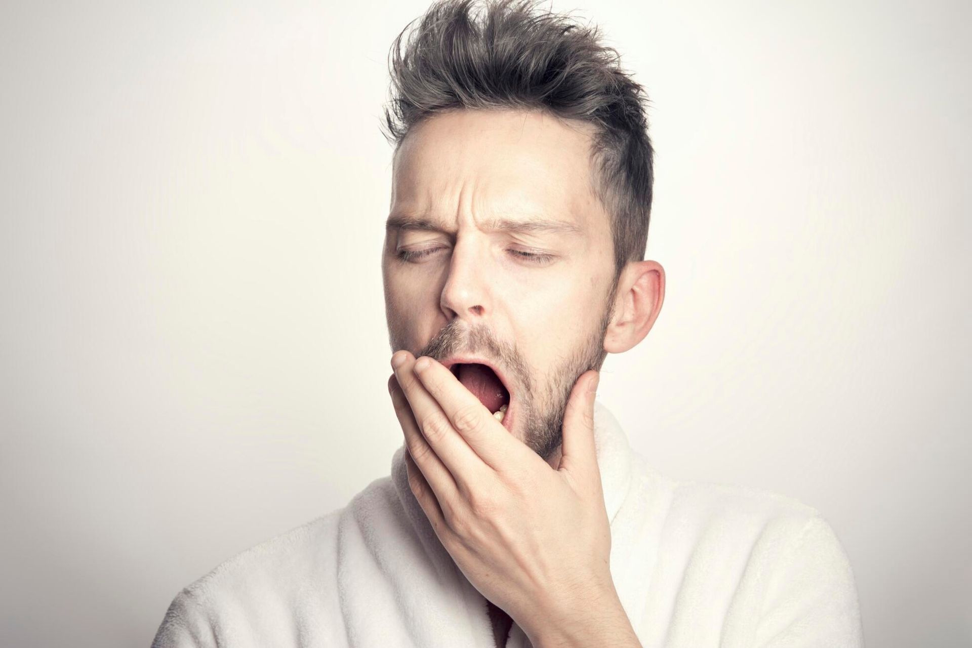 A person in a white robe yawns with their hand covering their mouth against a plain background.