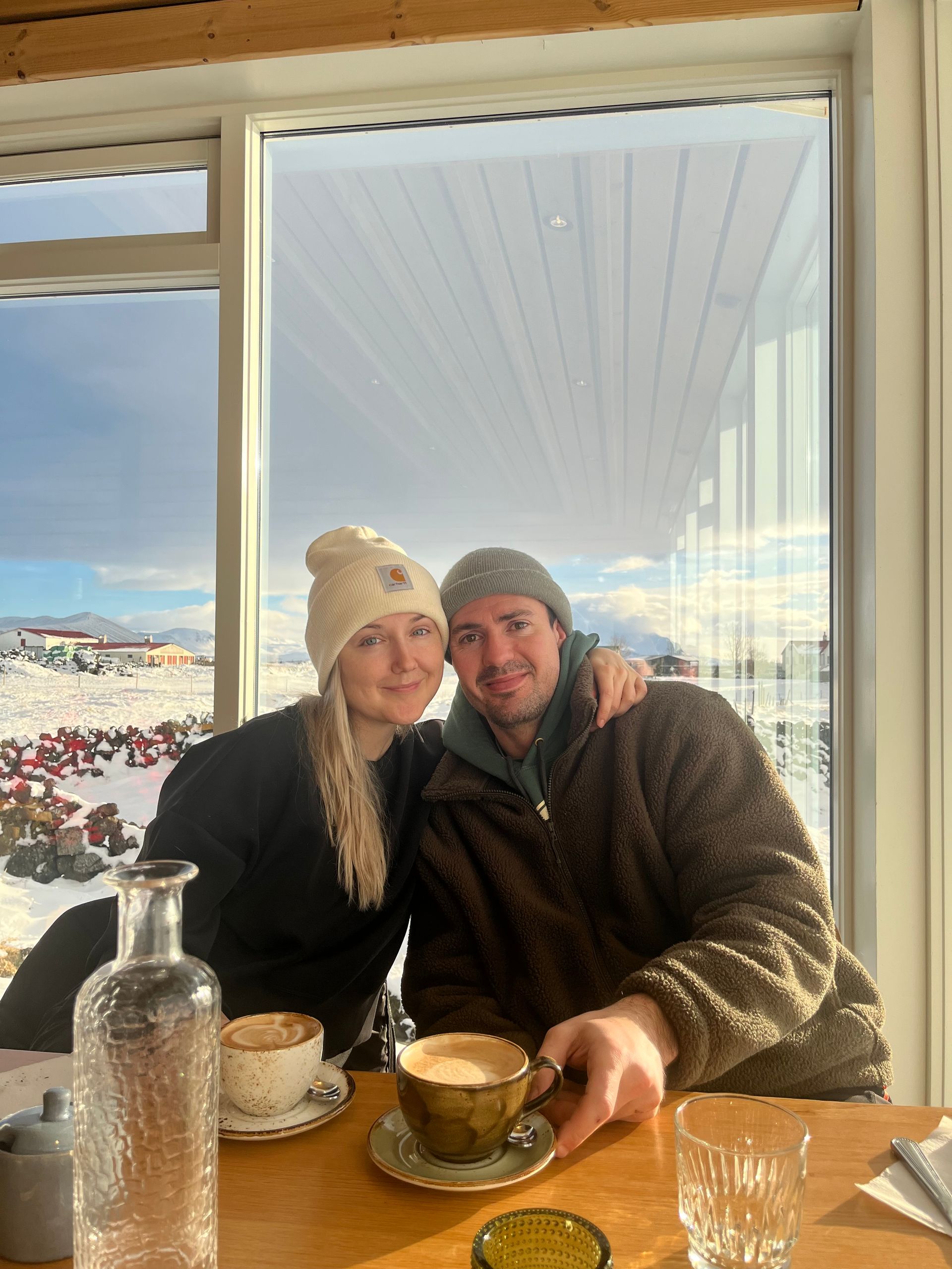 Couple seated at a cafe table by a window, smiling with cups of coffee and a snowy mountain view behind them