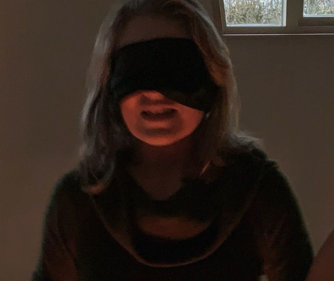 Person with a blindfold, illuminated by light, grimacing slightly, indoors.