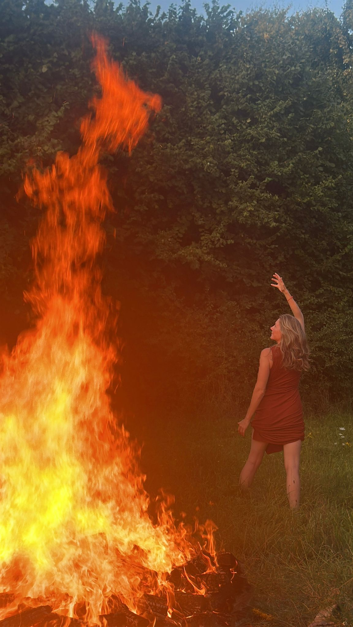 Woman in red dress near large bonfire, raising arm, with tall flames.