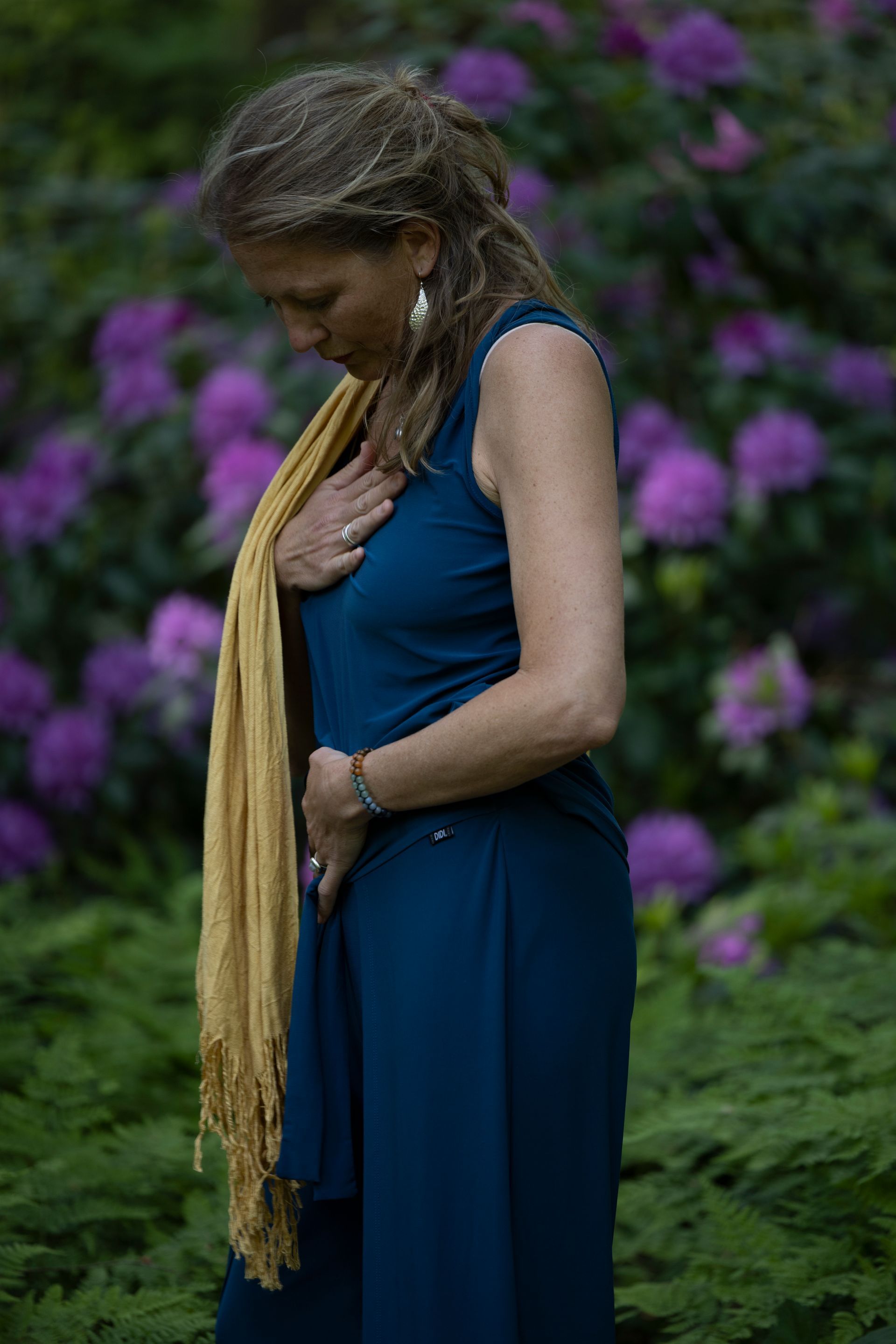 Een vrouw in een blauwe jurk en een gele sjaal staat voor paarse bloemen.