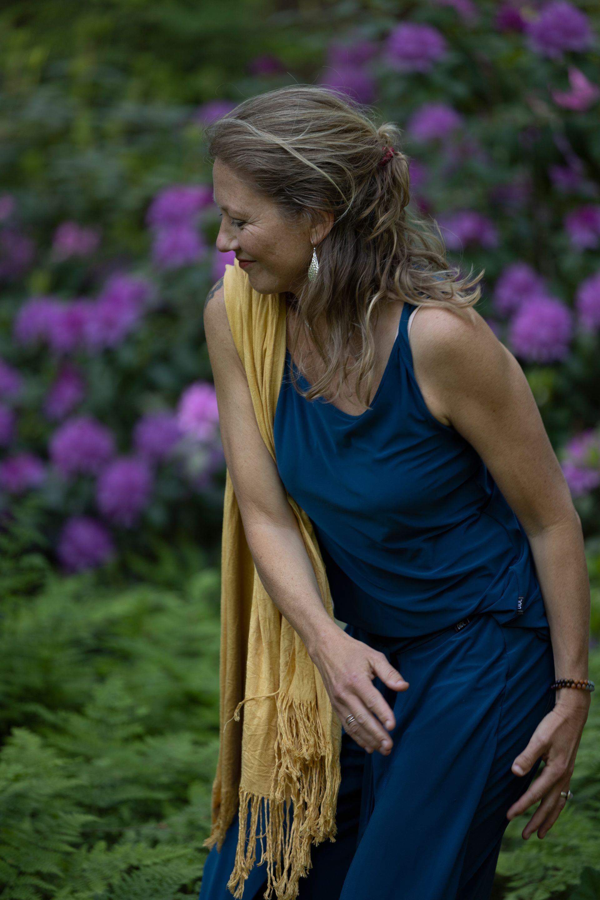 Een vrouw in een blauwe jurk en een gele sjaal staat voor paarse bloemen.