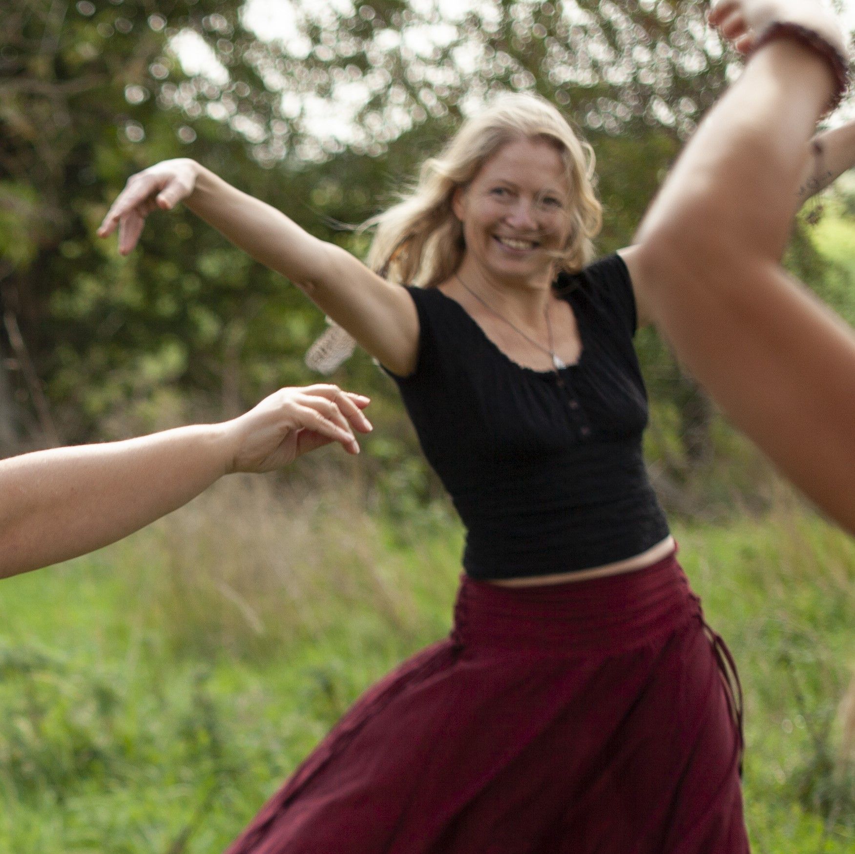Een vrouw danst buiten met uitgestrekte armen, gekleed in een rode rok en een zwarte top, omringd door anderen.