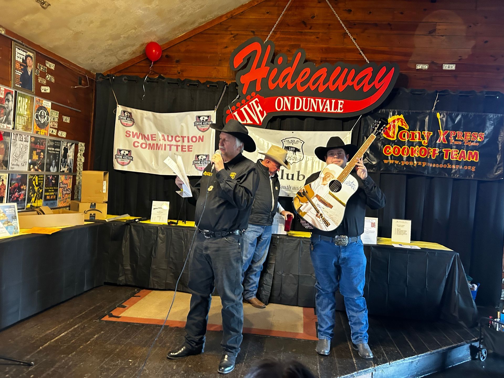 Three people on a stage at Hideaway on Dunvale, one speaking into a microphone, another holding a guitar, and a banner.