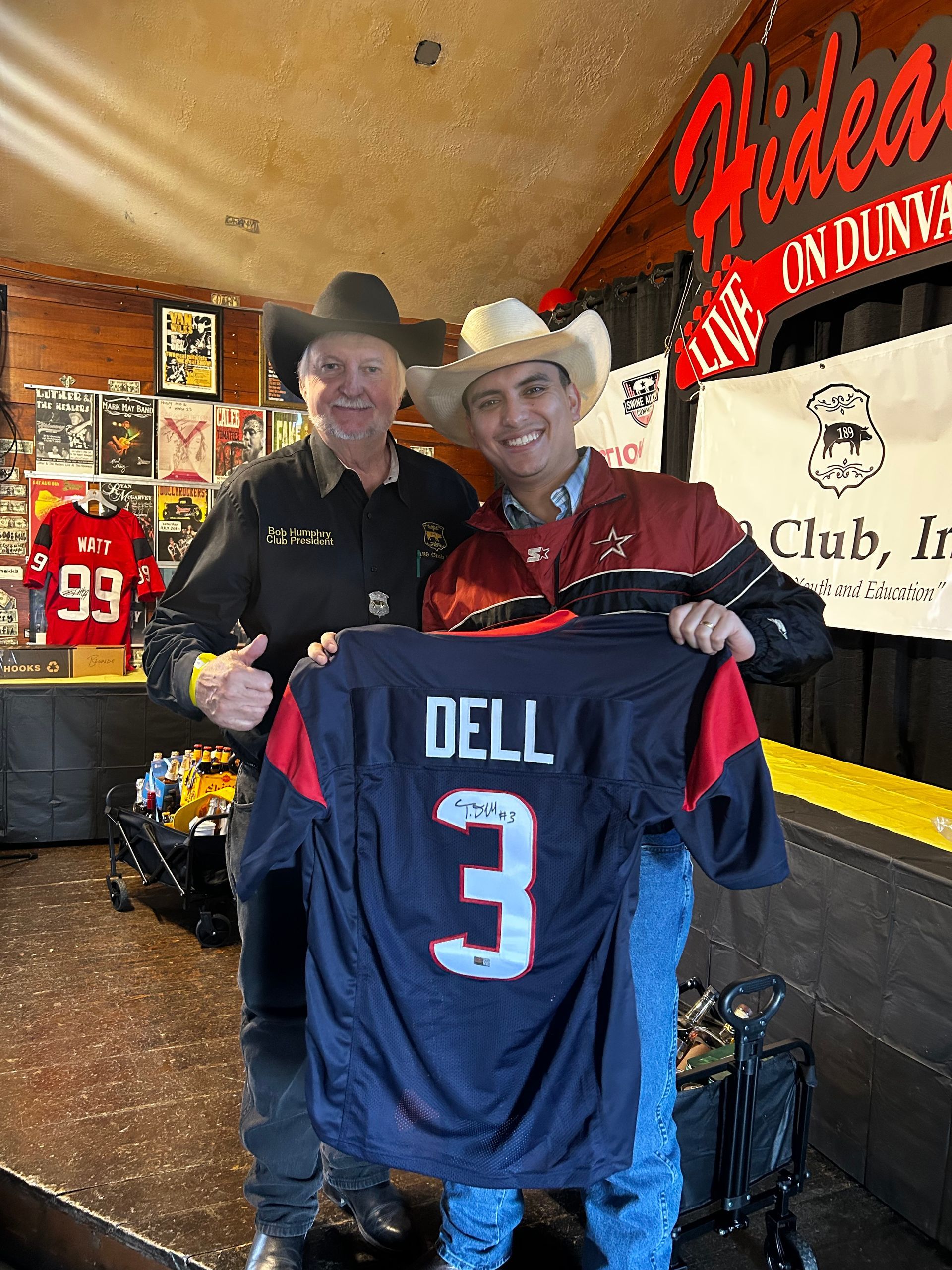 Two men in cowboy hats smile while holding up a dark navy Houston Texans jersey with