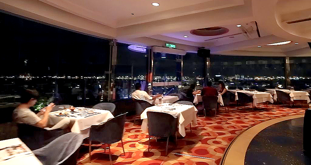 A dining room with tables, chairs, and patterned carpets, featuring large windows overlooking a city skyline at night.