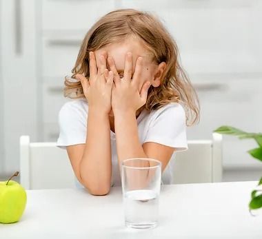 A little girl is sitting at a table covering her face with her hands.