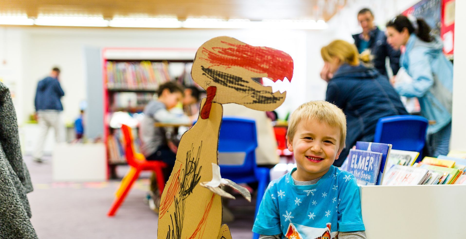 A boy with blonde hair wearing a blue t-shirt smiles next to a large cardboard cutout of a dinosaur. Behind them are people and books on shelves.