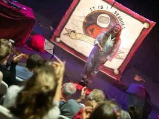 A woman in white overalls stands smiling on stage in front of an audience. A banner behind her says It Is I, Seagull.